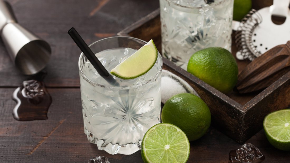 Gimlet Kamikaze cocktail in crystal glasses with lime slice and ice on wooden background with fresh limes and jigger with strainer.